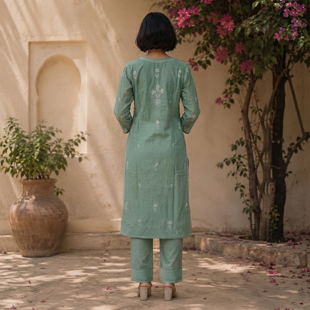 A woman viewed from behind wearing a sage green kurta and matching trousers with white embroidery, standing in a courtyard with beige walls, a large pot, and pink flowers.