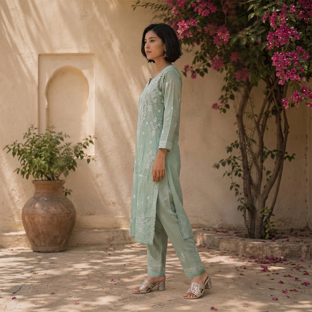 A woman models a light sage green three-piece ethnic suit with floral embroidery, standing in a sunlit courtyard with a beige wall, potted plants, and a flowering tree in the background.
