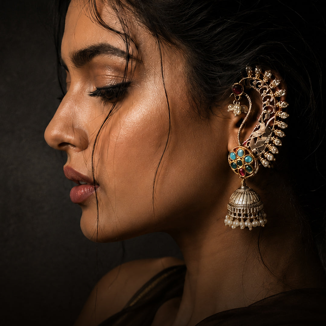 A close-up side profile of a woman wearing an ornate gold-toned traditional Indian jhumka and lobe hoop earring set with turquoise, red, and white stones, against a dark background.