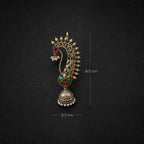 An ornate gold-toned peacock pendant with multicolored enamel details and a dangling bell, shown on a dark textured background with dimension markers indicating 8.5 cm height and 3.