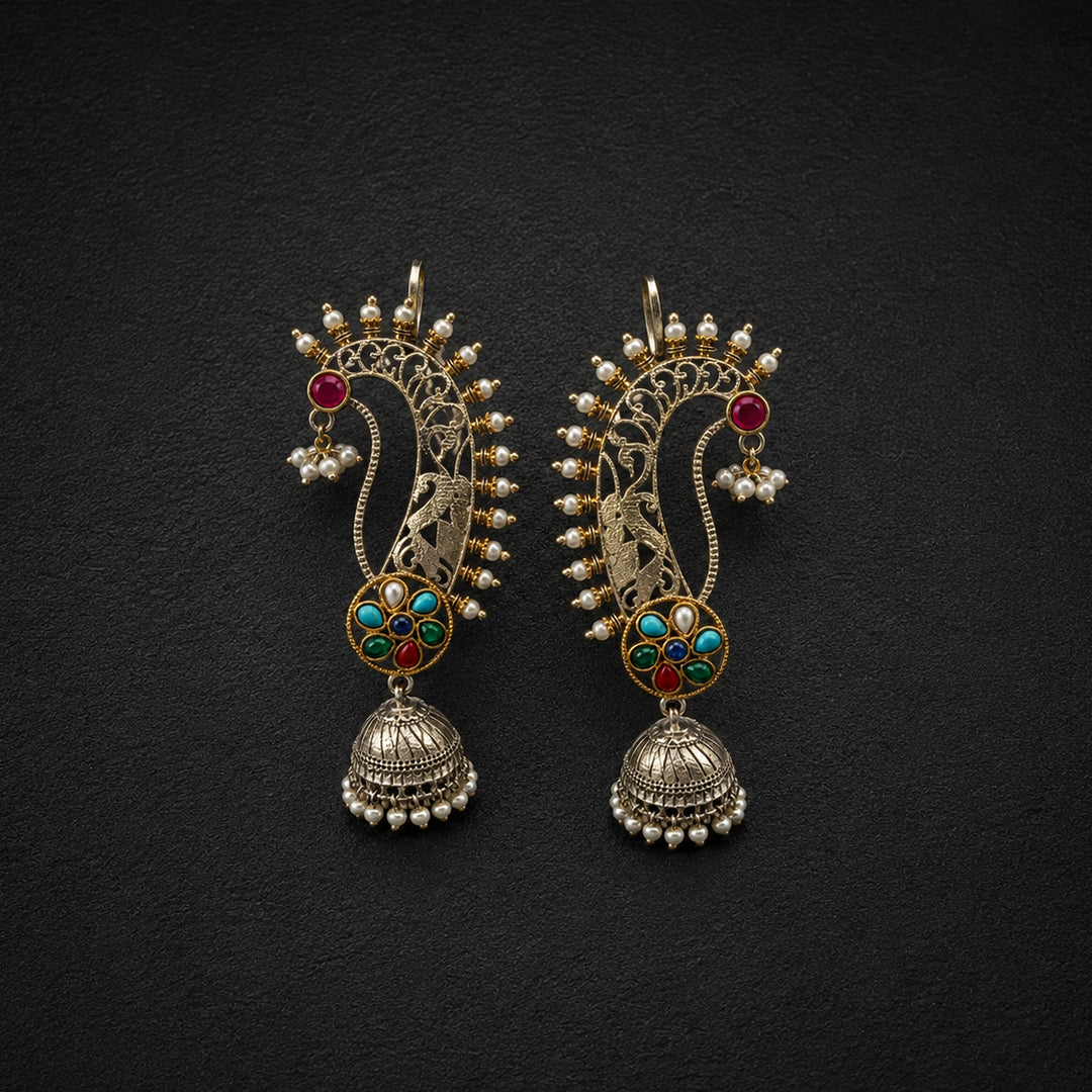 A pair of ornate Indian Jhumka earrings in gold-toned metal with red, green, and turquoise enamel details, set against a dark textured background.