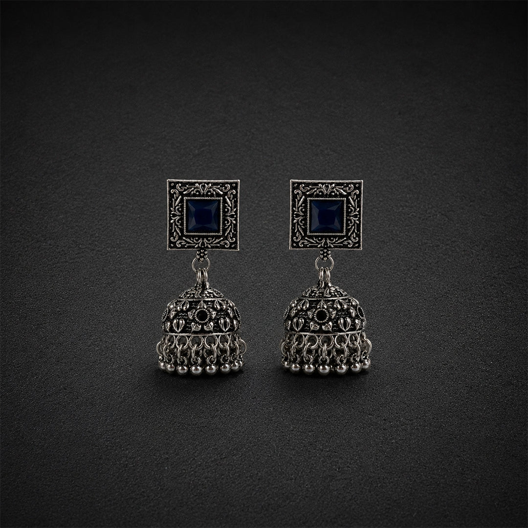 A pair of traditional Indian silver-toned jhumka earrings with dark blue square stones, displayed on a dark textured background.