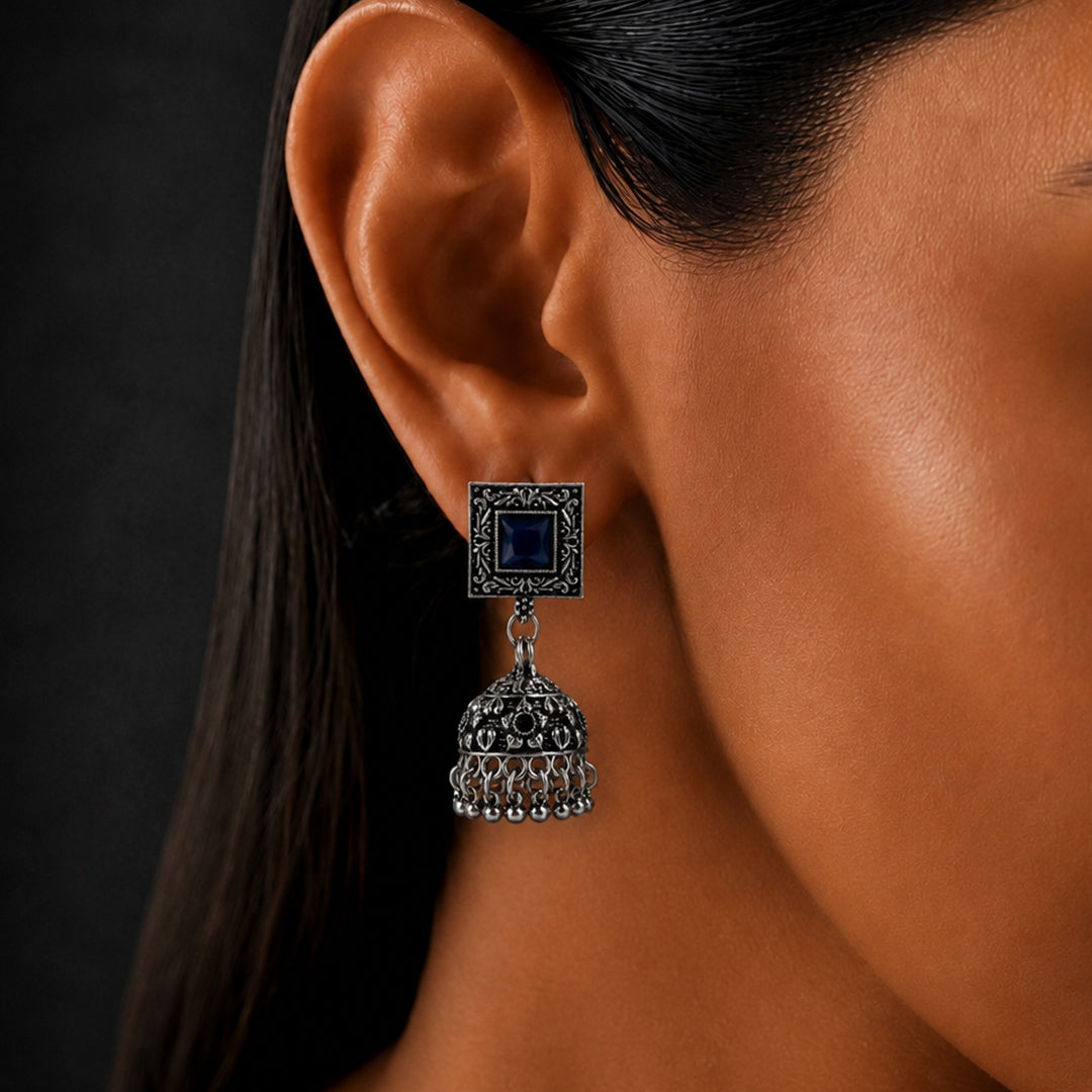 A close-up of a woman's ear wearing a silver-toned jhumka earring with a dark blue square stone and intricate filigree detailing.
