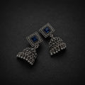 A pair of silver-toned jhumka earrings with square blue stone studs and ornate filigree bell-shaped dangling charms, displayed on a dark textured background.