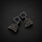 A pair of silver-toned jhumka earrings with square blue stone studs and ornate filigree bell-shaped dangling charms, displayed on a dark textured background.