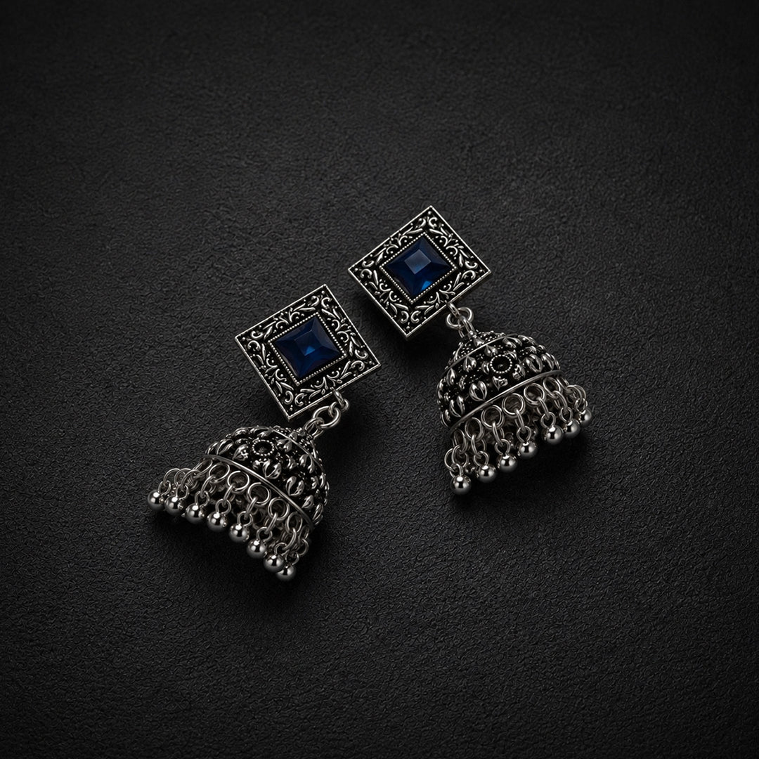 A pair of silver-toned jhumka earrings with square blue stone studs and ornate filigree bell-shaped dangling charms, displayed on a dark textured background.