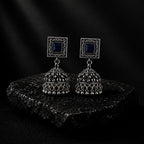 A pair of silver-toned Indian jhumka earrings with square blue stone studs, displayed on a dark textured stone slab against a dark background.