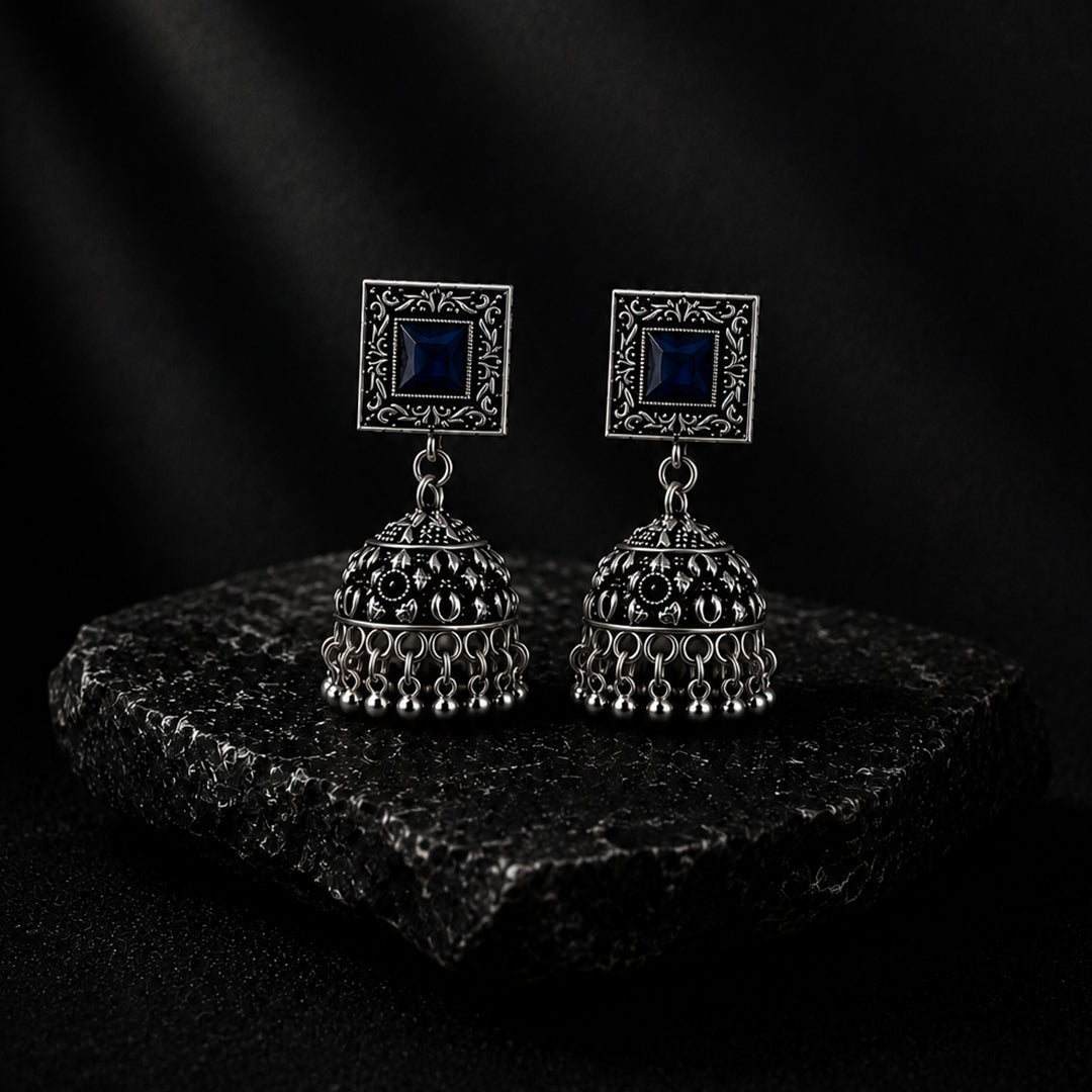 A pair of silver-toned Indian jhumka earrings with square blue stone studs, displayed on a dark textured stone slab against a dark background.