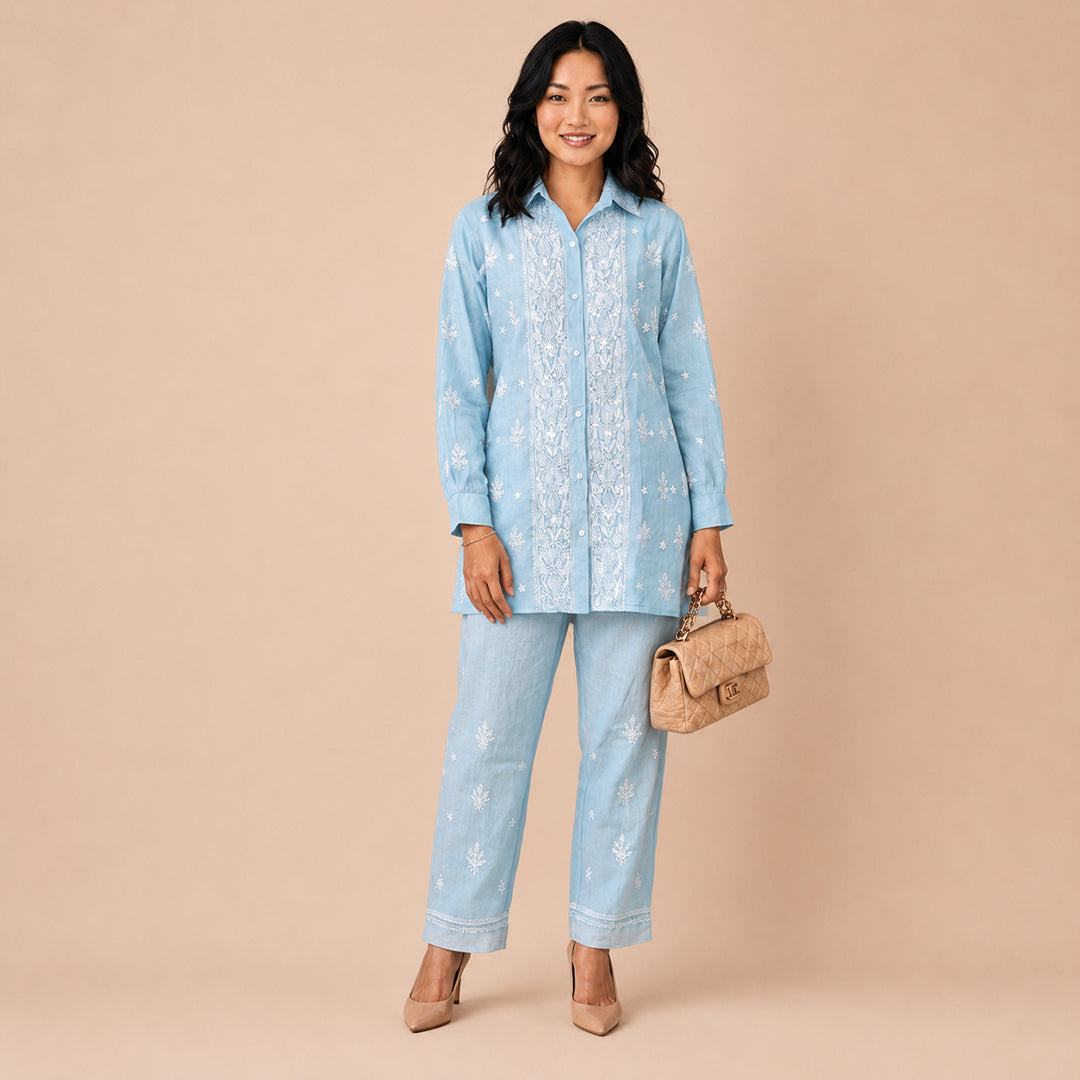 A woman modeling a light blue embroidered tunic and matching wide-leg pants set, holding a small tan handbag against a solid beige studio background.