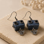A pair of dangling earrings with rectangular dark blue textured beads featuring black circular details, each hanging from a silver fishhook ear wire with a small silver bee charm below, displayed on a beige stone slab with dried flowers in the background.