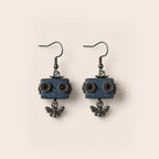 A pair of dangling earrings featuring rectangular blue wooden blocks with carved circular details resembling eyes, hanging from dark metal fishhook wires, with small dangling bee charms below each block, set against a solid light beige background.