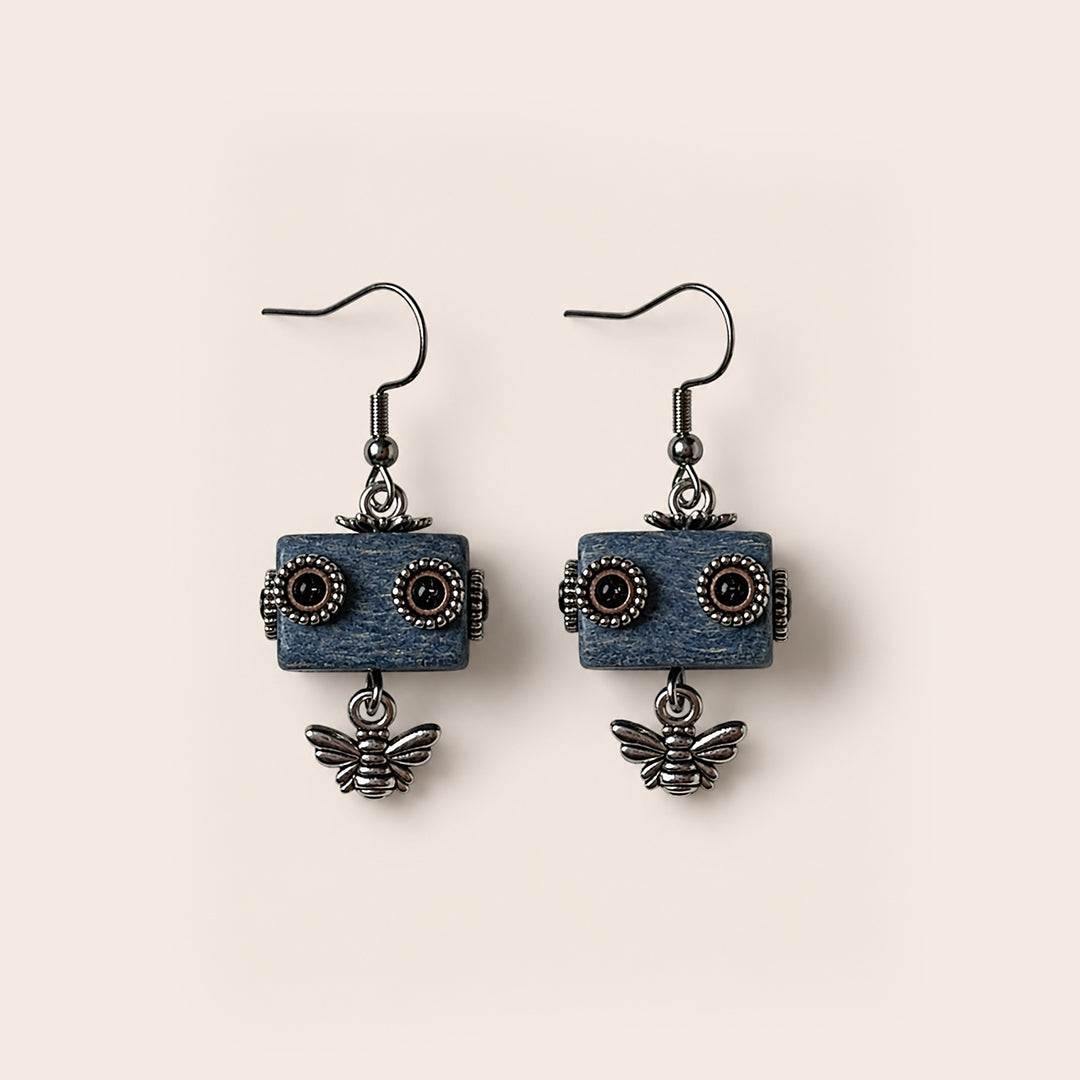 A pair of dangling earrings featuring rectangular blue wooden blocks with carved circular details resembling eyes, hanging from dark metal fishhook wires, with small dangling bee charms below each block, set against a solid light beige background.