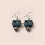 A pair of dangling earrings with rectangular blue wooden robot faces and small silver bee charms below, photographed on a plain warm beige background.