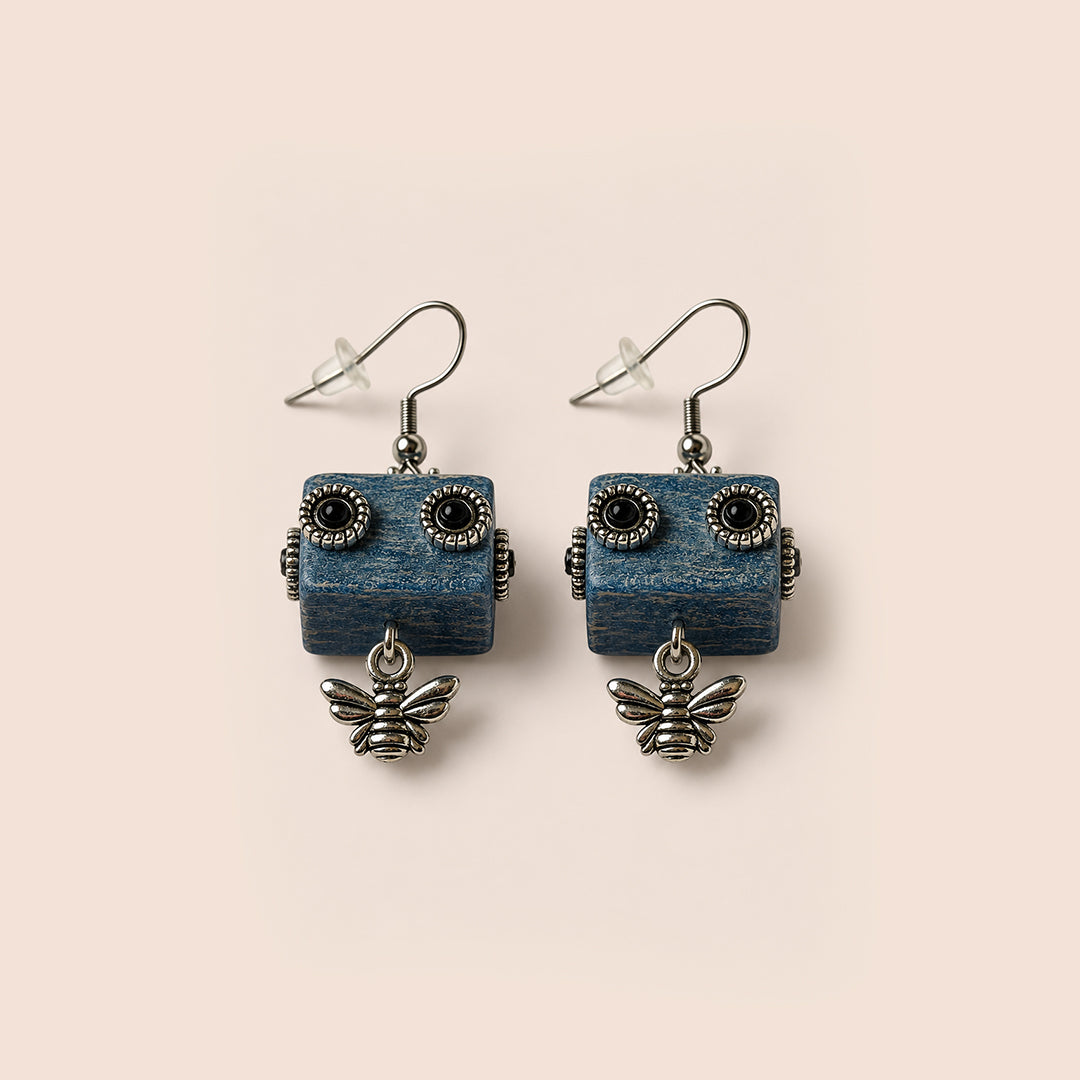 A pair of dangling earrings with rectangular blue wooden robot faces and small silver bee charms below, photographed on a plain warm beige background.