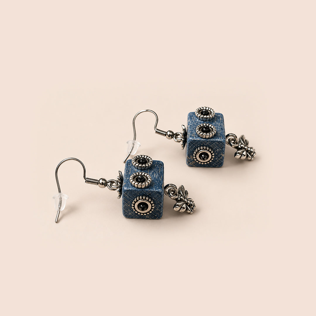 A pair of bohemian-style drop earrings featuring textured blue cube charms with black and gold eye details, hanging from silver-toned French hooks with small silver floral charms, set against a solid pale peach background.