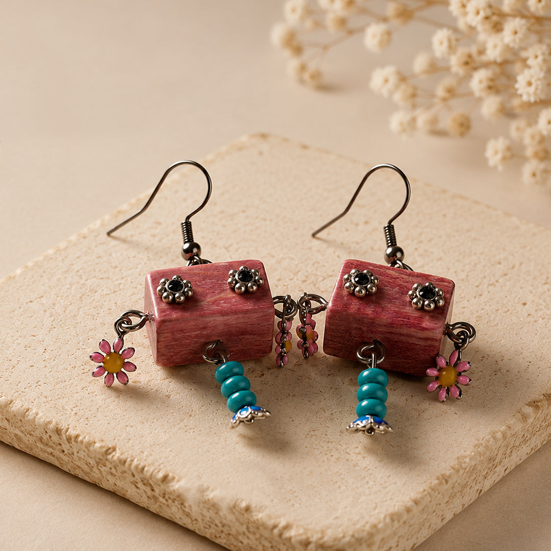 A pair of handmade dangle earrings displayed on a textured beige stone slab. The earrings feature rectangular pinkish-red wooden beads with silver floral studs, turquoise blue beads, and small pink daisy charms.