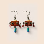 A pair of handmade dangle earrings featuring rectangular reddish-brown wooden blocks with metal flower charms, small pink flowers, and turquoise bead drops, set against a solid pale beige background.