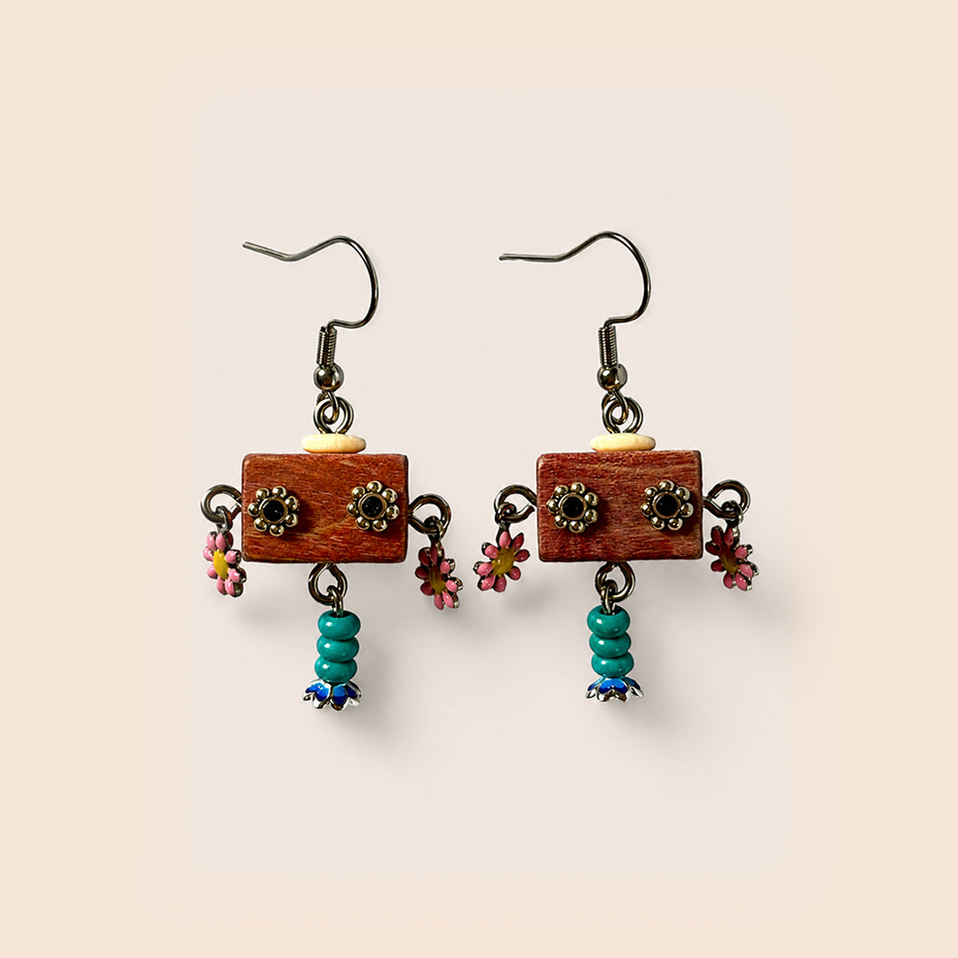 A pair of handmade dangle earrings featuring rectangular reddish-brown wooden blocks with metal flower charms, small pink flowers, and turquoise bead drops, set against a solid pale beige background.