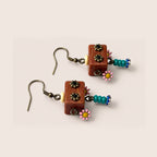 A pair of handmade dangle earrings featuring rectangular reddish-brown wooden cubes with three black floral charms, a small cream bead, a turquoise beaded section, and a pink and yellow daisy, shown on a light beige background.