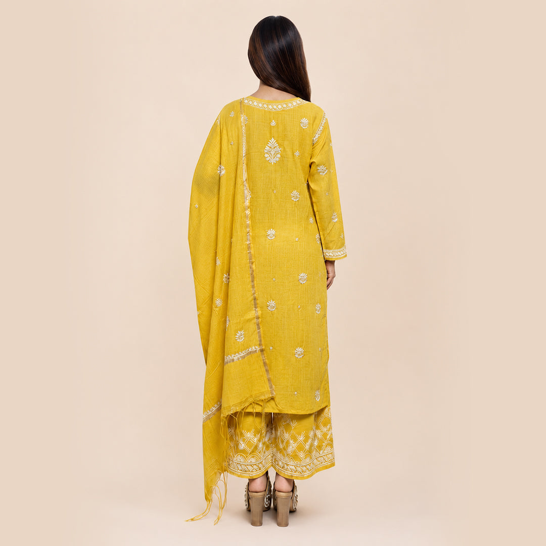 A woman shown from the back wearing a yellow traditional outfit with white embroidered details and a matching dupatta, set against a solid beige background.