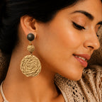 A close-up of a woman wearing a large dangle earring with a dark round stud and a textured beige woven spiral drop, shown against her ear and neck.