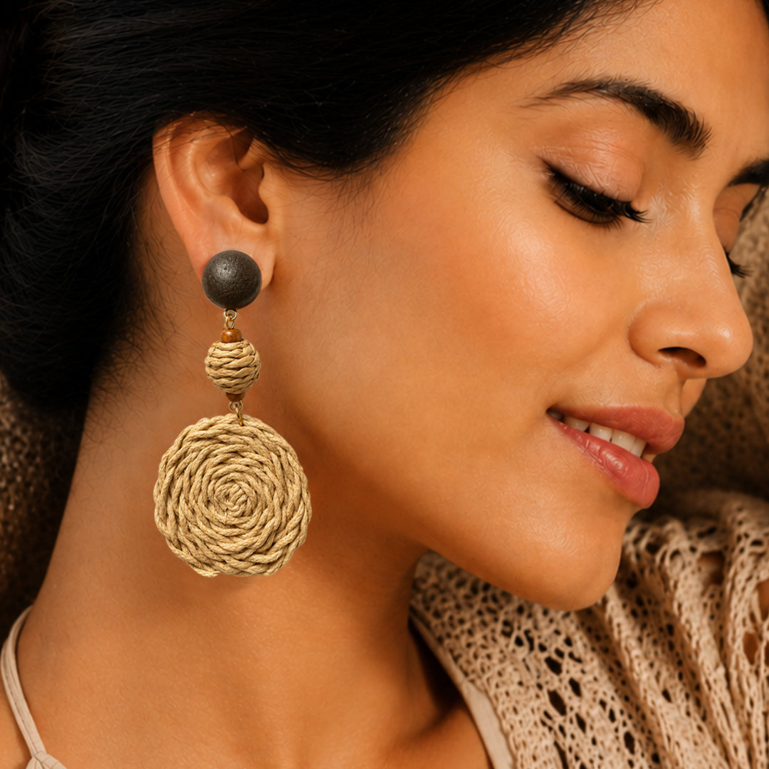 A close-up of a woman wearing a large dangle earring with a dark round stud and a textured beige woven spiral drop, shown against her ear and neck.
