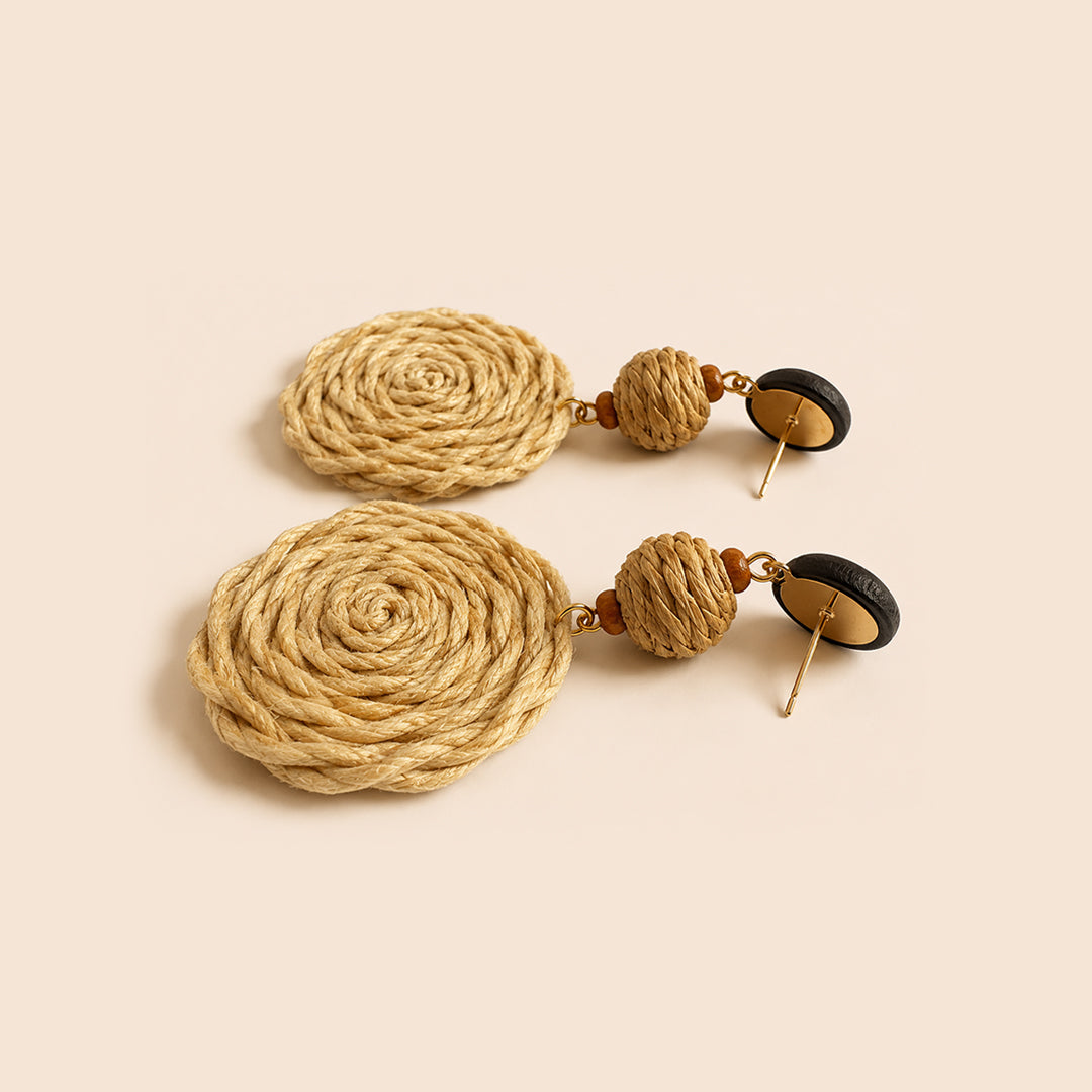 A pair of bohemian-style dangle earrings featuring large circular rosettes wrapped in natural jute rope, with small textured beads, gold-toned metal connectors, and round black-and-gold wooden studs, displayed on a solid light beige background.