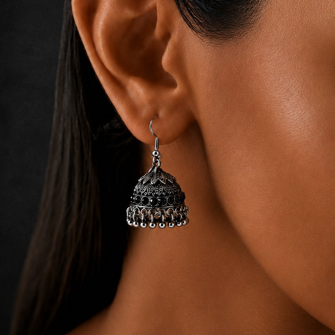 A close-up of a woman's ear wearing a detailed silver-toned jhumka earring with ornate filigree and dangling bead elements, set against a dark background.