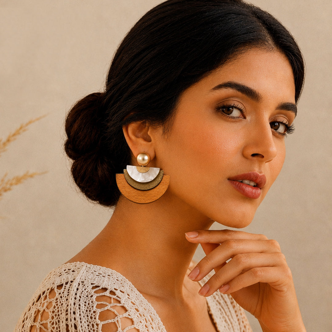 A woman with dark hair in a bun wears a pair of tiered semicircle earrings with a gold-toned top disc, a white shell-like middle disc, and a light brown wooden bottom disc, paired with a small gold pearl stud.