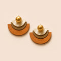 A pair of geometric semi-circular earrings with a wooden half-moon base, a textured metallic gold crescent, a white shell-like petal, and a small gold sphere on a warm beige background.