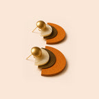 A pair of geometric stud earrings featuring a gold ball stud, a white shell disc, a dark brown textured disc, and a warm orange-brown wooden crescent shape, displayed on a solid pale peach background.