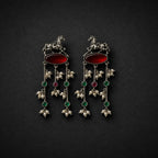 A pair of ornate silver-toned dangle earrings with red oval stones, green and red round stones, and white pearl-like floral drops, displayed on a dark textured background.