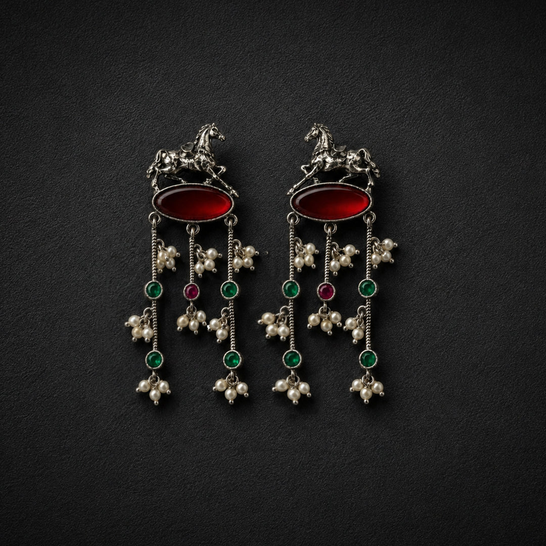 A pair of ornate silver-toned dangle earrings with red oval stones, green and red round stones, and white pearl-like floral drops, displayed on a dark textured background.