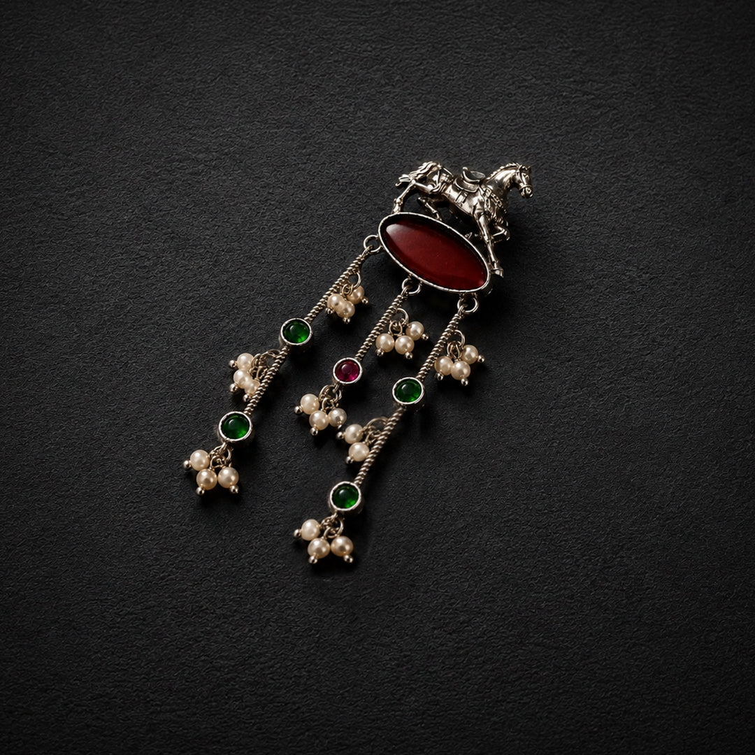 A silver-toned traditional Indian pendant featuring a horse motif, an oval red cabochon stone, and three dangling chains with green and red stones and pearl-like beads, displayed on a dark textured background.
