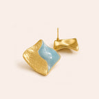 A pair of organic-shaped gold and light blue earrings with textured gold surfaces and smooth blue inlays, displayed on a warm off-white background.