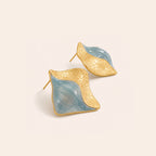 A pair of organic-shaped gold and light blue earrings with textured gold surfaces and smooth blue inlays, displayed on a warm off-white background.