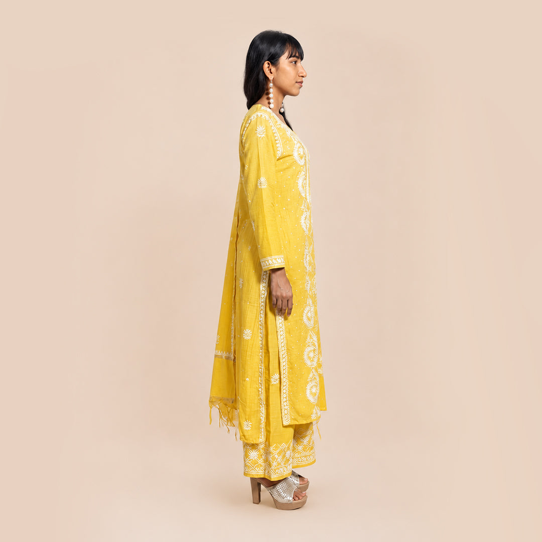 A woman shown in profile wearing a bright yellow traditional outfit with white embroidery, standing against a plain beige studio background.