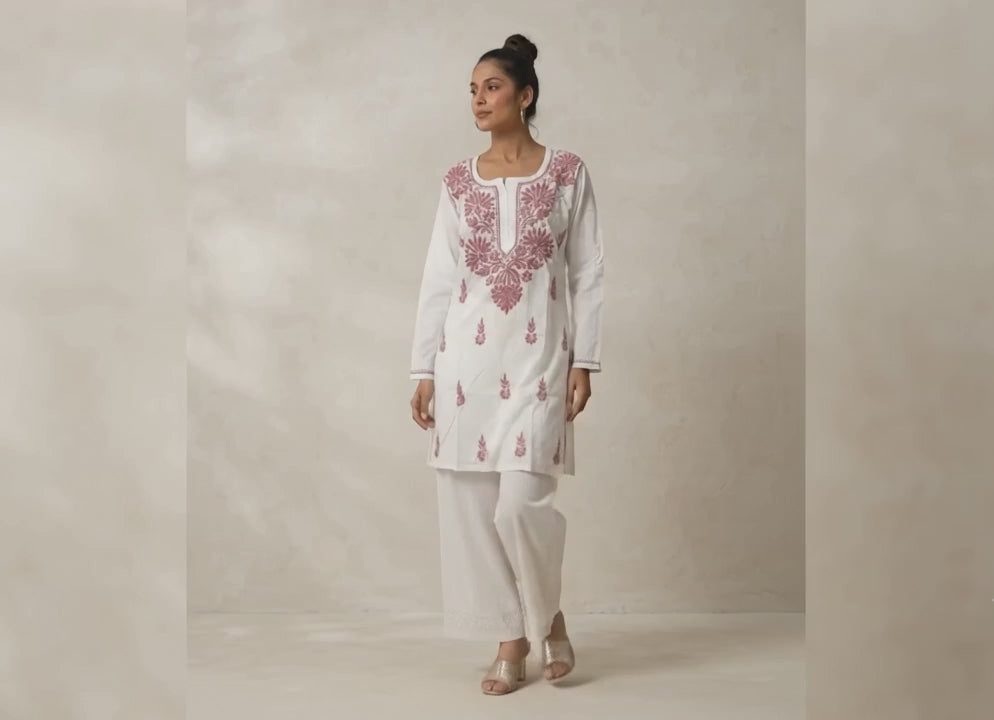 A woman in profile wearing a white traditional outfit with pink floral embroidery, paired with white pants and metallic heeled sandals, set against a textured beige wall with soft leaf shadows.